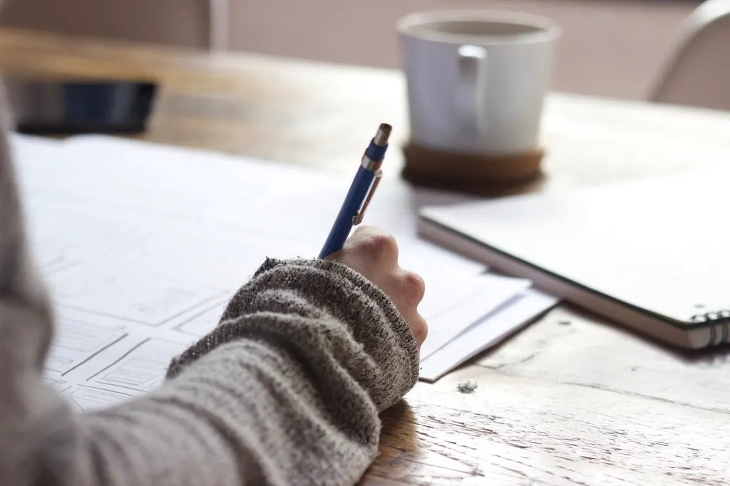 A person in a knitted jumper writes on papers at a wooden table with a mug.