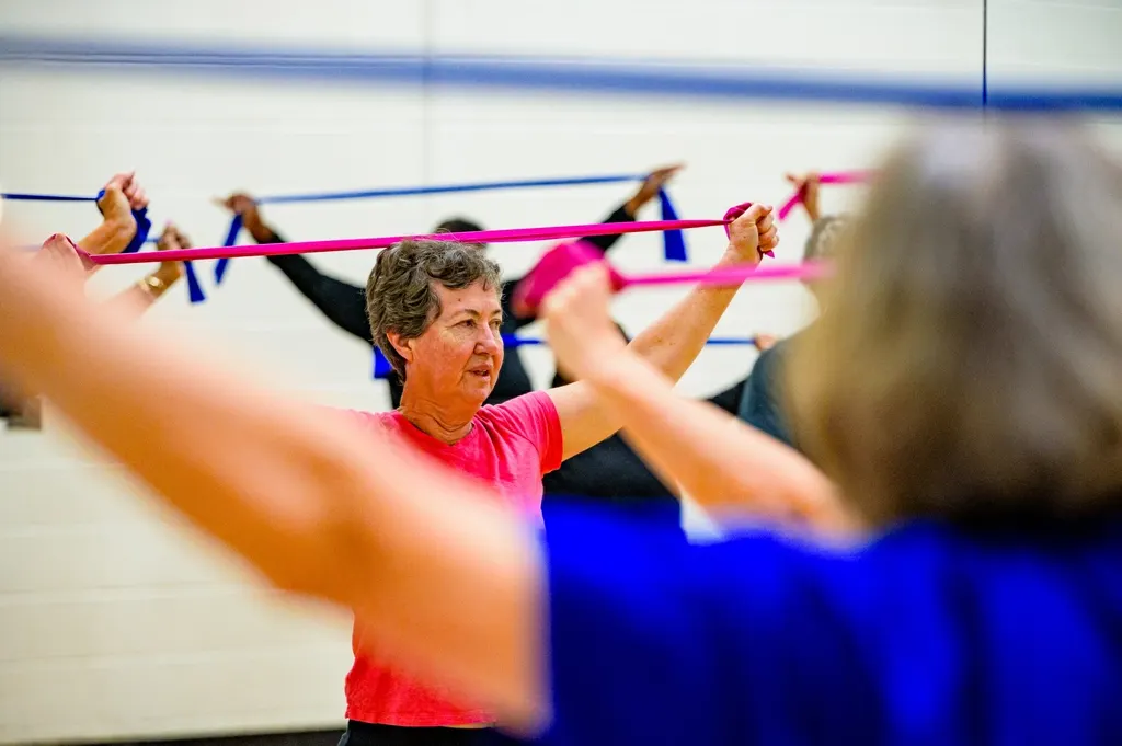 An older woman stretches a resistance band overhead in a group fitness class.