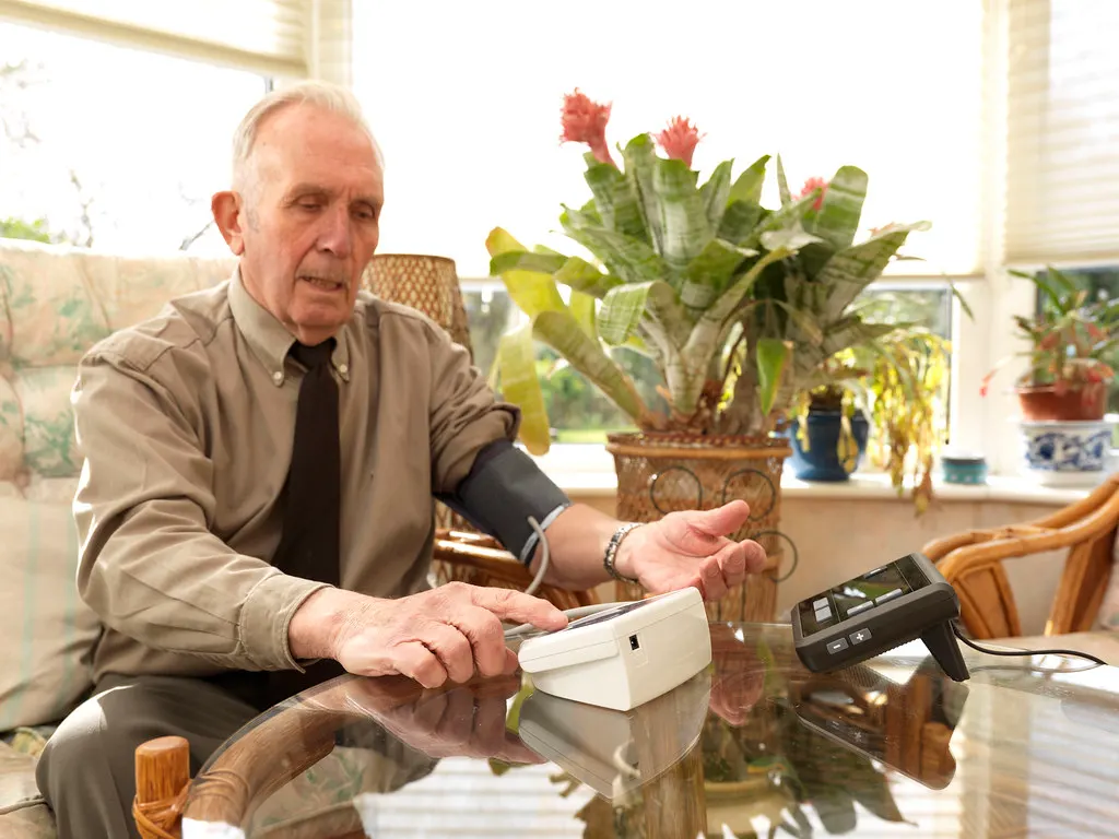 An older man checks his own blood pressure at a table in a sunny conservatory.