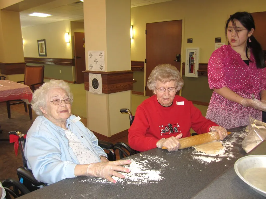 Two older women in wheelchairs roll out pastry at a baking session with a helper.