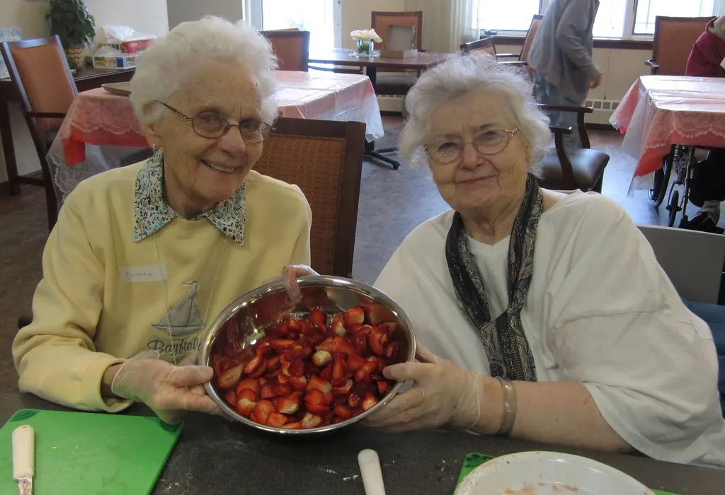 Two smiling older women hold up a bowl of freshly cut strawberries.