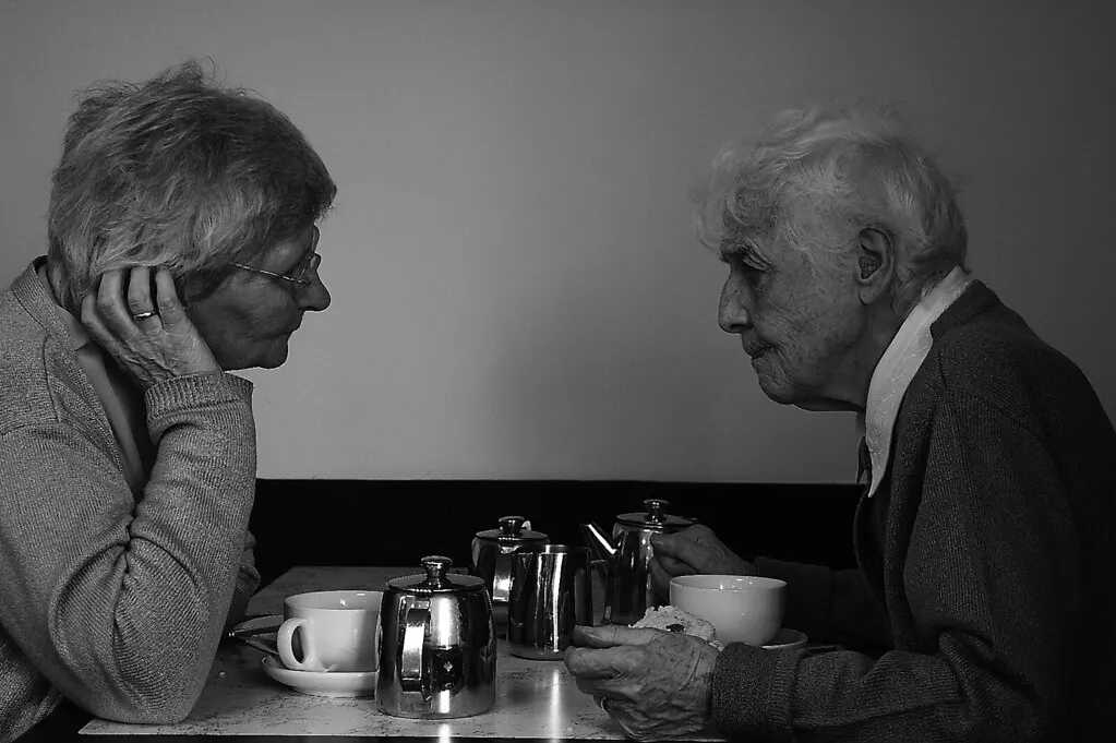Two older people chat over tea and a scone at a café table, in black and white.