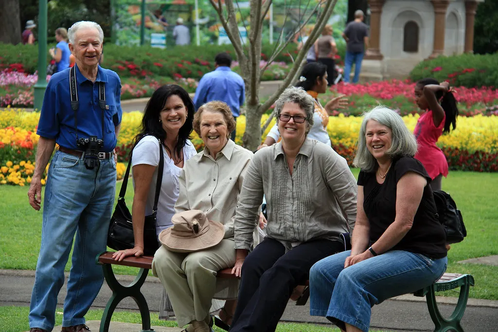 A smiling group of older friends sit on a bench in a colourful flower garden.