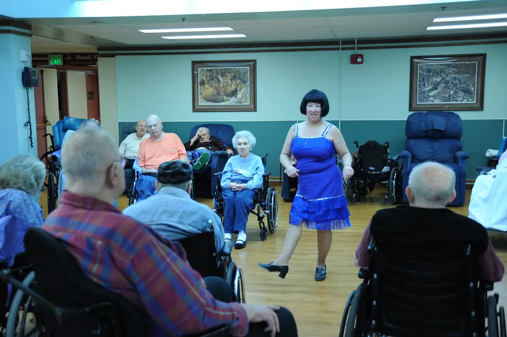 A performer in a blue dress entertains residents in a care home day room.