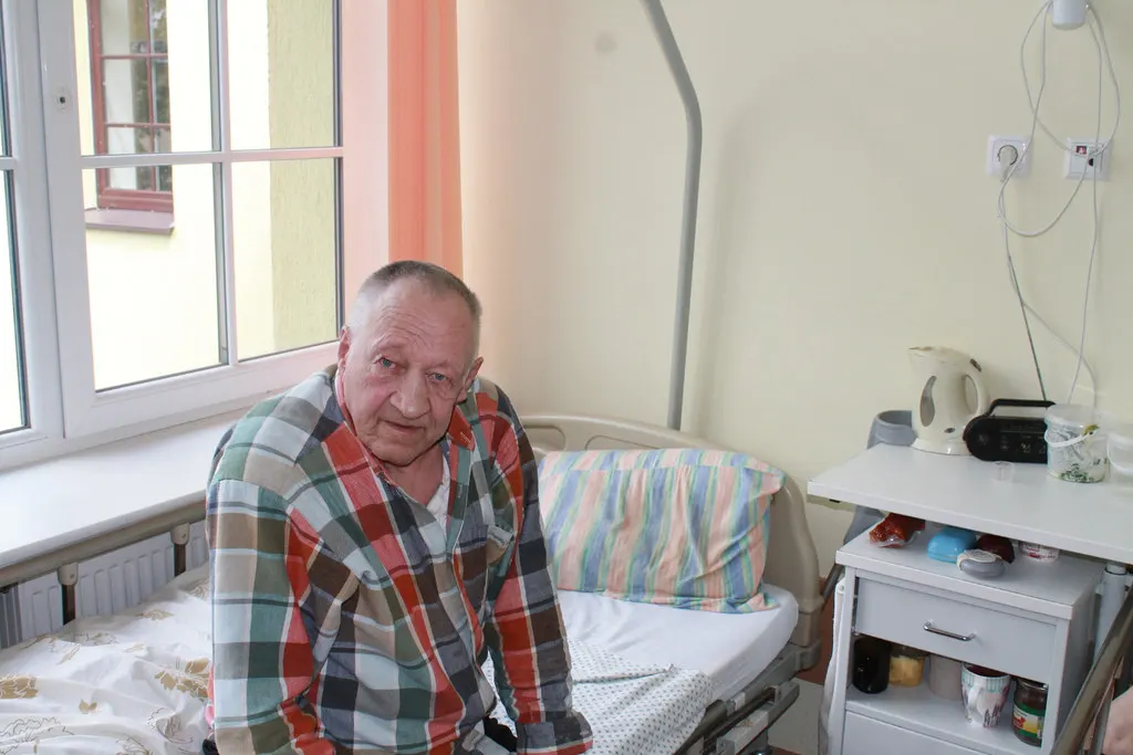 An older man in a checked shirt sits on the edge of his bed in a care room.