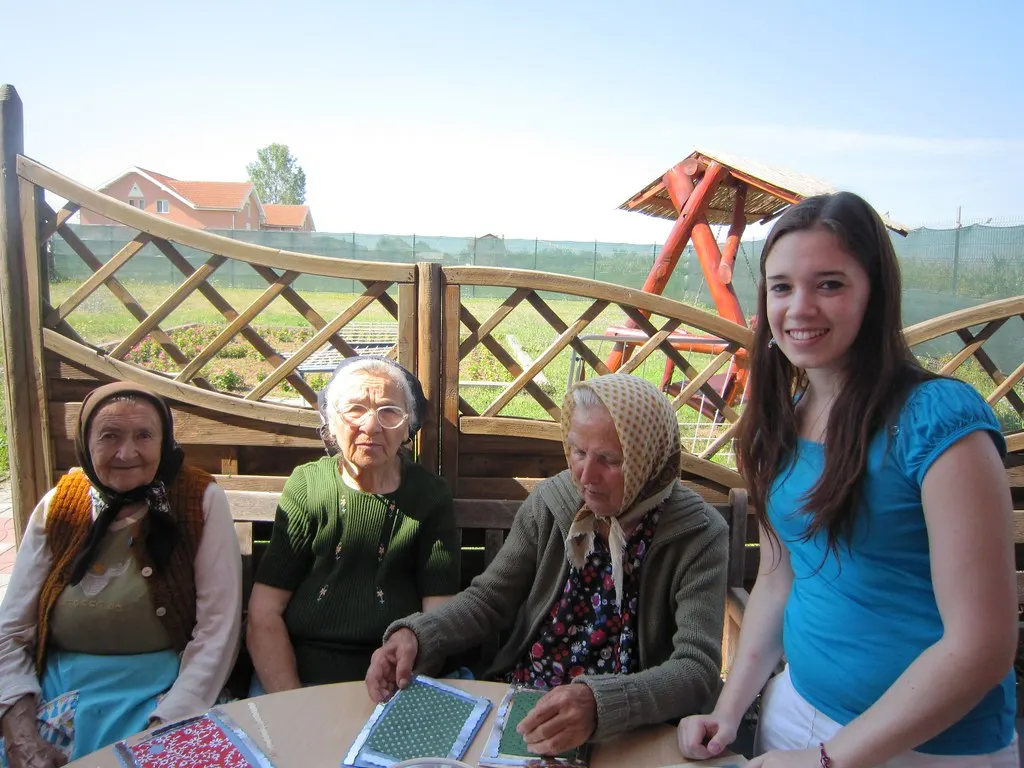 A smiling young volunteer sits with three older women doing a craft activity outdoors.