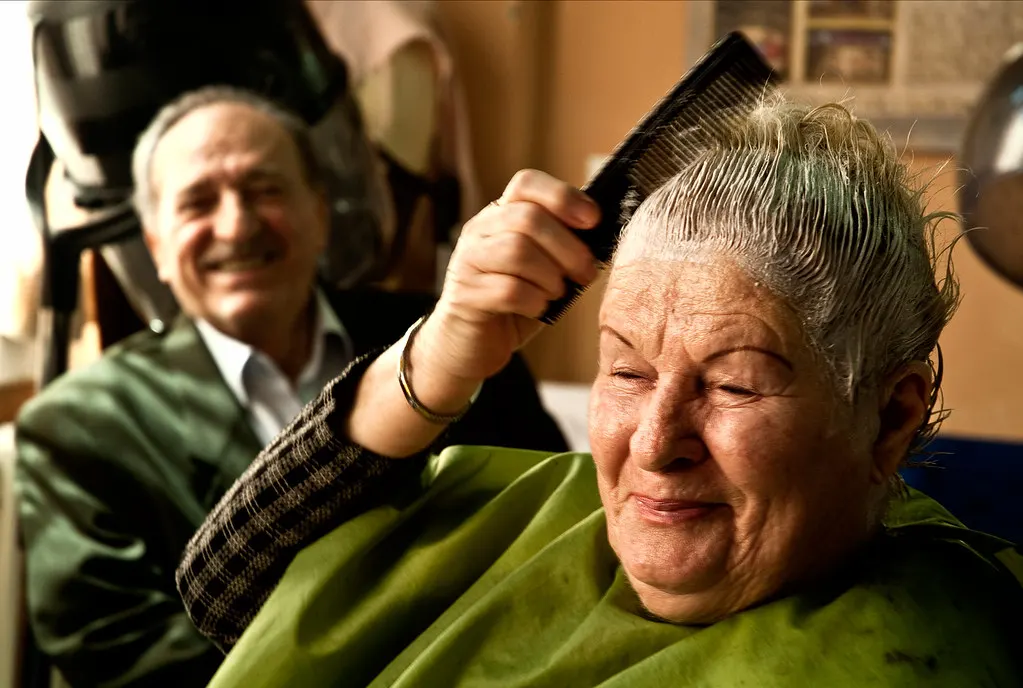 A smiling older woman combs her freshly washed hair at a hairdresser, a man laughing behind her.